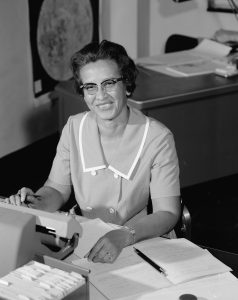 Katherine Johnson, seated at a desk, surrounded by papers. A poster of the moon is on the wall behind her. 
