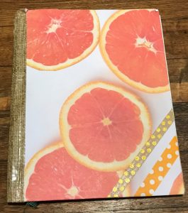 notebook decorated with a piece of scrapbook paper with photos of grapefruits and washi tape stripes in the lower corner: polka dot natural color with gold and white polka dot on orange. The binding is duct tape. 