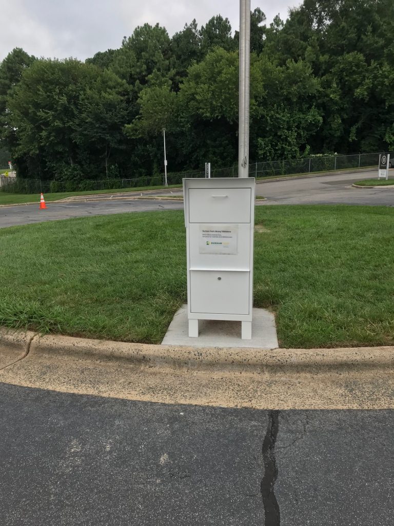 White outdoor drive-up book drop located in Parking Lot C with flagpole in background